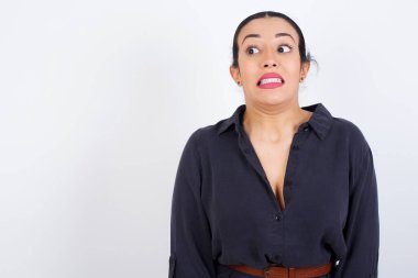 Tüh! Beyaz stüdyo arka planına karşı gri elbise giyen genç ve güzel bir Arap kadının portresi dişlerini sıkar ve şaşırtıcı bir şekilde yana bakar.,