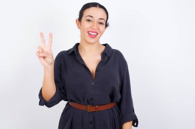 Beyaz stüdyo arka planına karşı gri elbise giyen genç ve güzel bir Arap kadın iki numaralı parmaklarıyla gülümseyerek kendinden emin ve mutlu bir şekilde onu işaret ediyor..
