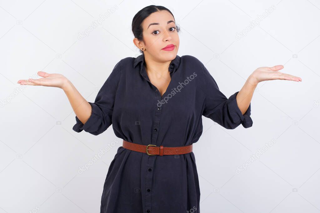 Puzzled and clueless Arab woman wearing dress against white studio background with arms out, shrugging shoulders, saying: who cares, so what, I don't know. Negative human emotions.