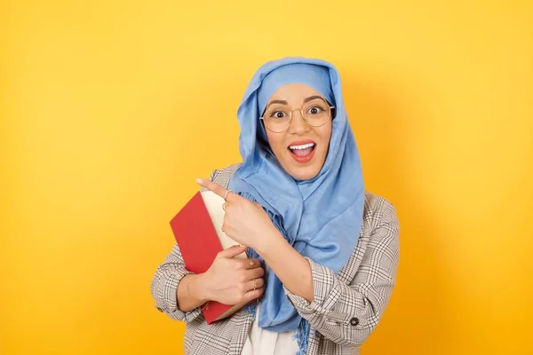 看那边 年轻美丽的穆斯林女子头戴头巾 拿着书本 一边指着你 一边微笑着 一边站在工作室的背景下 — 图库照片