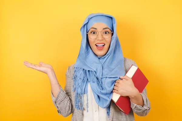 身穿头巾的积极乐观的年轻穆斯林妇女说 多么令人兴奋啊 有着令人惊奇的表情 在空旷的空间里用张开的手在孤立的黄色背景上展示了一些东西 广告概念 — 图库照片