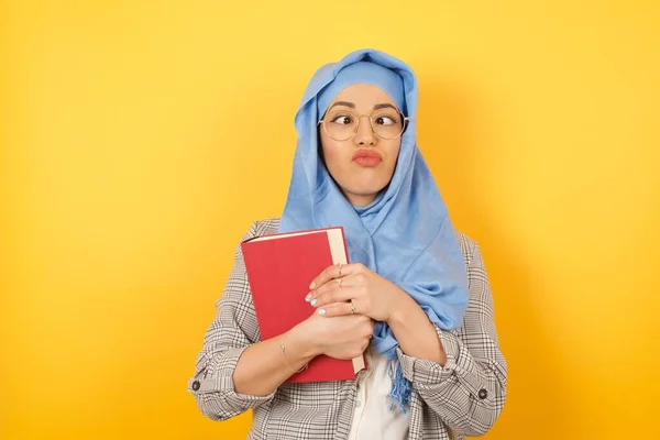 Funny comic muslim woman wearing hijab crosses eyes, puts lips, makes grimace, foolishness after all day studying. Clueless woman nerd with awkward expression has fun alone, plays fool.