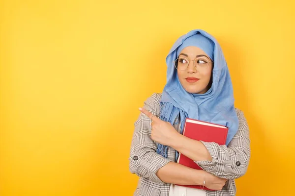 年轻漂亮的穆斯林女人 头戴头巾 拿着书 摆出棘手的样子 摆出姿势站在旁边 摆出一副相机的样子 女设计师用食指展示可爱的产品 Gesture概念 — 图库照片