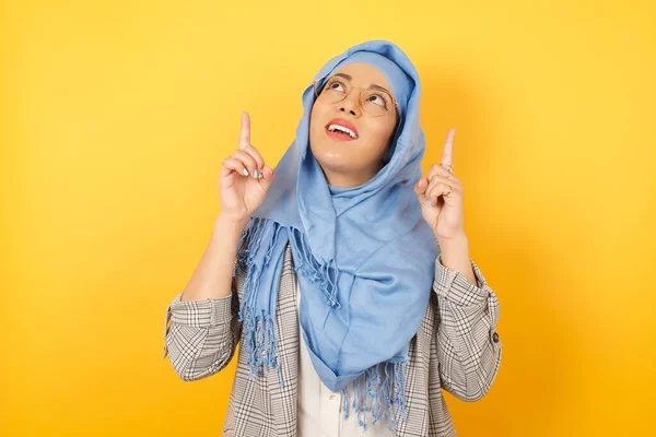 Horizontal shot of successful friendly looking young muslim woman wearing hijabexclaiming excitedly, pointing both index fingers up, indicating something shocking on blank copy space wall.