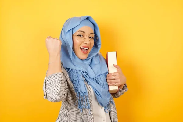喜形于色的年轻穆斯林女子头戴头巾 头戴头巾 喜气洋洋地捧着书本 积极地从幸福中得到消息 握紧拳头 做出胜利的姿态 反应概念 — 图库照片