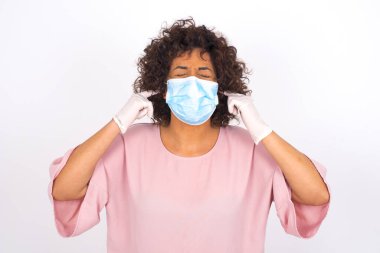 Yüzü maskeli genç bir kadın kulaklarını kapatıyor. Beyaz arka planda izole edilmiş. Bulaşıcı hastalıklara karşı koruma. Stüdyoda poz veren günlük kıyafetler giyen genç bir kadın.