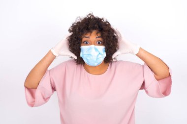 Yüz maskeli genç kadın beyaz arka planda soyutlanmış bir şekilde şoka uğruyor. Bulaşıcı hastalıklara karşı koruma. Stüdyoda poz veren günlük kıyafetler giyen genç bir kadın.