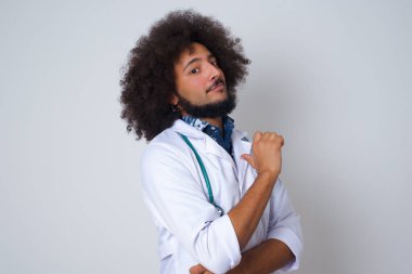 Neşeli, genç Afrikalı Amerikalı doktor neşeli, memnun ve kendinden emin görünüyor. Başparmağıyla işaret ediyor. Gri stüdyo arka planında izole edilmiş..