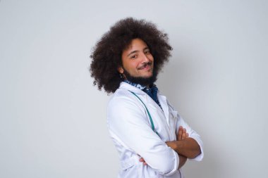 Beyaz önlüklü neşeli genç bir doktorun kolları çaprazlanmış bir şekilde içeride duruşu. Kameraya bakıp gülümsüyor. Kendine güvenen kız..