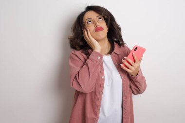 Akıllı telefonlu genç ve güzel bir kadın stüdyoya bakıyor. Beyaz duvara karşı cep telefonu tutan gözlüklü ve pembe ceketli bir Arap kadın.