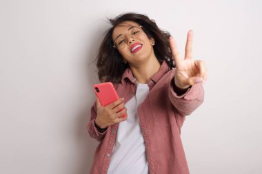 Genç, güzel, esmer, Arap kadın. Stüdyoda zafer jesti içeren pembe tişörtlü, akıllı telefonlu.