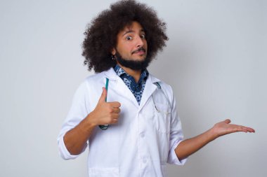 Genç, mutlu, doktor üniforması giyen, başparmağını kaldıran, diğer elini de evin içinde gösteren bir doktor. İyi iş çıkardın. Sağlık üniforması giyiyor..