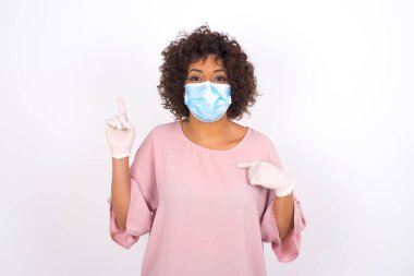 Yüzü maskeli genç bir kadın beyaz arka planda izole edilmiş. Bulaşıcı hastalıklara karşı koruma. Stüdyoda poz veren günlük kıyafetler giyen genç bir kadın.