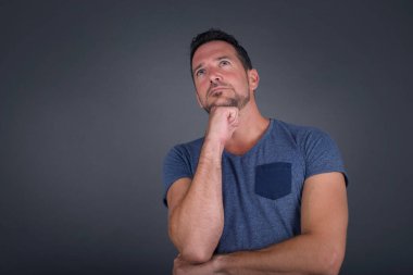 Düşünceli, gülümseyen, beyaz tişört giyen genç bir adamın portresi elini çenesinin altında tutuyor, yan bakıyor, ilgi çekici bir şey düşünüyor ya da merak ediyor, tesadüfen giyinmiş, gri stüdyoya karşı poz veriyor. Karar alma kavramı.