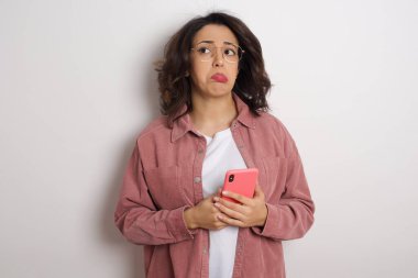 Akıllı telefonlu genç ve güzel bir kadın stüdyoda üzgün. Beyaz duvara karşı cep telefonu tutan gözlüklü ve pembe ceketli bir Arap kadın.