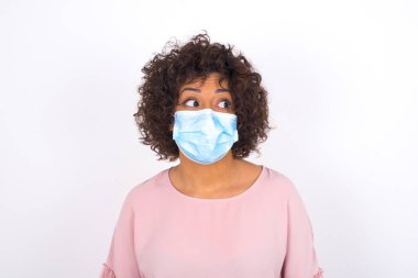 Yüzü maskeli genç bir kadın beyaz arka planda soyutlanmış bir şekilde yüzünü çeviriyor. Bulaşıcı hastalıklara karşı koruma. Stüdyoda poz veren günlük kıyafetler giyen genç bir kadın.