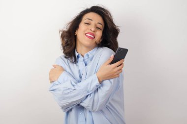 Stüdyoda mavi gömlekli, beyaz duvarlı, akıllı telefonlu genç bir iş kadını.