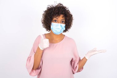 Beyaz arka planda, baş parmağı yukarıda, maskeli genç bir kadın. Günlük kıyafetler giymiş, yüz maskesi takan genç bir kadın. Bulaşıcı hastalıklara karşı koruma.