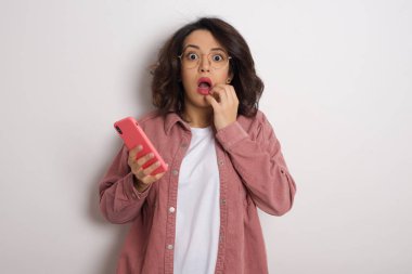 Akıllı telefonlu genç ve güzel bir kadın stüdyoda şok geçiriyor. Beyaz duvara karşı cep telefonu tutan gözlüklü ve pembe ceketli bir Arap kadın.