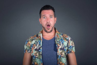 Ağzı açık duygusal çekici erkek, büyük bir şaşkınlık ve korkuyu ifade eder, beyaz beton arka planda poz verir, kameraya bakar. Beklenmedik haberler ve insani tepkiler.