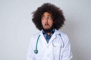  Genç doktor züppe ifadeli, buruşuk dudaklı, kaşlarını kaldıran şüpheli ve şüpheci bir ifadeyle şüpheli bir ifadeyle bakıyor. Gri arka planda kapalı duruyor..