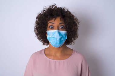 Beyaz arka planda soyutlanmış yüz maskeli genç bir kadın şok geçirdi. Bulaşıcı hastalıklara karşı koruma. Stüdyoda poz veren günlük kıyafetler giyen genç bir kadın.