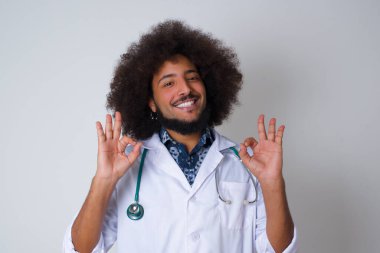 Tıp üniforması giyen çekici genç bir doktorun iki elinde de onay işareti olması sevindirici bir ifadeyle iyimser olması güzel. Gri duvara yaslanarak.