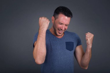  Beyaz bir adam tişört giyip başarısını ve zaferini kutluyor. Yumruklarını zevkten dört köşe ediyor. Saçlı şanslı adam hedefine ve hedeflerine ulaştığına seviniyor. Olumlu duygular, duygular.