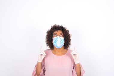 Yüzü maskeli genç bir kadın beyaz arka planda izole bir şekilde yukarı bakıyor. Bulaşıcı hastalıklara karşı koruma. Stüdyoda poz veren günlük kıyafetler giyen genç bir kadın.