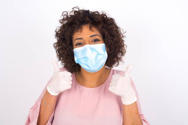 Beyaz arka planda, baş parmakları yukarıda, maskeli genç bir kadın. Günlük kıyafetler giymiş, yüz maskesi takan genç bir kadın. Bulaşıcı hastalıklara karşı koruma.