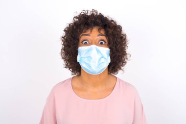 Yüz maskeli genç kadın beyaz arka planda soyutlanmış bir şekilde şoka uğruyor. Bulaşıcı hastalıklara karşı koruma. Gündelik kıyafetler giymiş genç bir kadın.