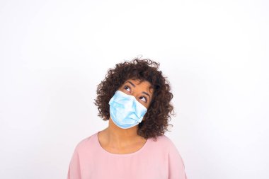 Yüzü maskeli genç bir kadın beyaz arka planda soyutlanmış bir şekilde yüzünü çeviriyor. Bulaşıcı hastalıklara karşı koruma. Stüdyoda poz veren günlük kıyafetler giyen genç bir kadın.