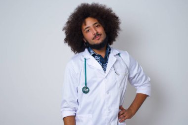 Neşeli Afrikalı Amerikalı doktor, elini kalçasında tutuyor, geniş bir şekilde gülümsüyor, arkadaşlarının doğum gününde hazırladığı sürprizi bekliyor, gri arka planda izole edilmiş..