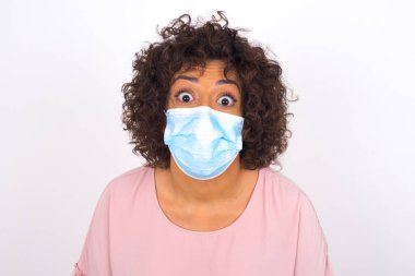 Yüz maskeli genç bir kadının beyaz arka planda kafası karışmış. Günlük kıyafetler giymiş, yüz maskesi takan genç bir kadın. Bulaşıcı hastalıklara karşı koruma.