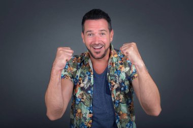 Gri arka planda Hawaii çiçekli gömlek giyen şık, yakışıklı beyaz erkek portresi. Gülümsüyor ve kazanan hareketi yapıyor. 