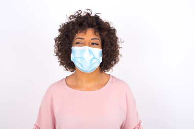 Yüzü maskeli genç bir kadın beyaz arka planda izole bir şekilde gülümsüyor. Günlük kıyafetler giymiş, yüz maskesi takan genç bir kadın. Bulaşıcı hastalıklara karşı koruma.