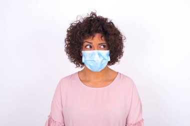 Yüzü maskeli genç bir kadın beyaz arka planda soyutlanmış bir şekilde yüzünü çeviriyor. Bulaşıcı hastalıklara karşı koruma. Stüdyoda poz veren günlük kıyafetler giyen genç bir kadın.