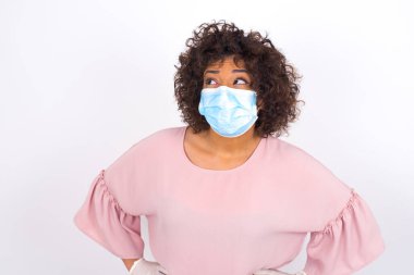 Yüzü maskeli genç bir kadın beyaz arka planda soyutlanmış bir şekilde yüzünü çeviriyor. Günlük kıyafetler giymiş, yüz maskesi takan genç bir kadın. Bulaşıcı hastalıklara karşı koruma.