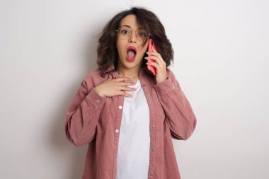 Stüdyoda akıllı telefondan konuşan kafası karışmış güzel bir kadın. Beyaz duvara karşı cep telefonu tutan gözlüklü ve pembe ceketli bir Arap kadın.