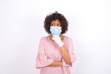 Maskeli genç bir kadın beyaz arka planda soyutlanmış olduğunu düşünüyor. Bulaşıcı hastalıklara karşı koruma. Stüdyoda poz veren günlük kıyafetler giyen genç bir kadın.