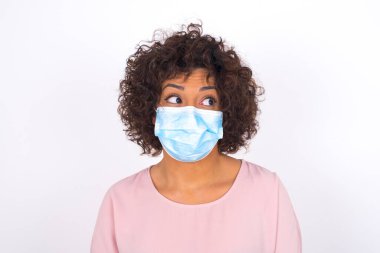 Yüzü maskeli genç bir kadın beyaz arka planda soyutlanmış bir şekilde yüzünü çeviriyor. Bulaşıcı hastalıklara karşı koruma. Stüdyoda poz veren günlük kıyafetler giyen genç bir kadın.