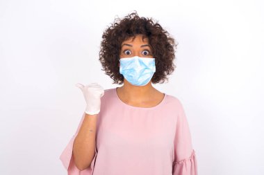 Yüz maskeli genç bir kadın beyaz arka planda izole edilmiş sırt çantaları gösteriyor. Bulaşıcı hastalıklara karşı koruma. Stüdyoda poz veren günlük kıyafetler giyen genç bir kadın.