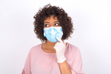 Maskeli genç bir kadın beyaz arka planda soyutlanmış olduğunu düşünüyor. Bulaşıcı hastalıklara karşı koruma. Stüdyoda poz veren günlük kıyafetler giyen genç bir kadın.