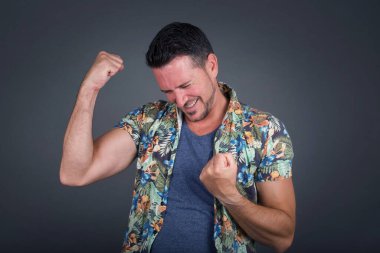 Gri arka planda Hawaii çiçekli gömlek giyen şık, yakışıklı beyaz erkek portresi. Gülümsüyor ve kazanan hareketi yapıyor. 