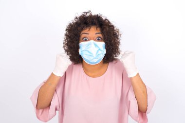 Beyaz arka planda yumruklarını gösteren maskeli genç bir kadın. Bulaşıcı hastalıklara karşı koruma. Stüdyoda poz veren günlük kıyafetler giyen genç bir kadın.