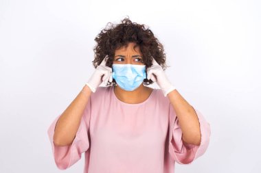 Beyaz arka planda başı ağrıyan maskeli genç bir kadın. Bulaşıcı hastalıklara karşı koruma. Stüdyoda poz veren günlük kıyafetler giyen genç bir kadın.