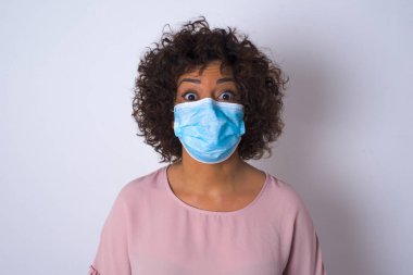 Beyaz arka planda maskeli, kafası karışmış genç bir kadın var. Bulaşıcı hastalıklara karşı koruma. Stüdyoda poz veren günlük kıyafetler giyen genç bir kadın.