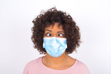 Yüzünde maske olan genç bir kadın beyaz arka planda izole edilmiş çılgın bir yüz yapıyor. Bulaşıcı hastalıklara karşı koruma. Stüdyoda poz veren günlük kıyafetler giyen genç bir kadın.