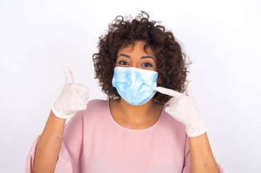 Beyaz arka planda, baş parmağı yukarıda, maskeli genç bir kadın. Bulaşıcı hastalıklara karşı koruma. Stüdyoda poz veren günlük kıyafetler giyen genç bir kadın.