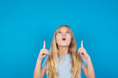 beautiful caucasian girl wearing blue t-shirt  being amazed and surprised looking and pointing up with fingers showing something strange. clipart